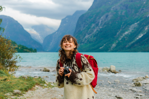 Young Caucasian woman in knitted sweater standing with vintage camera and photographing scenic views of the lake in Norway