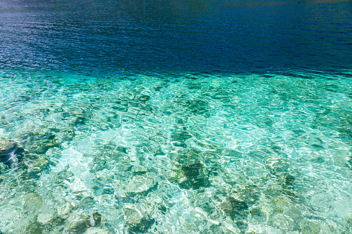Surface of the sea water as the background. The upper part of the water appears in a dark blue hue, while the lower part is transparent, providing a view of the sea bottom. The clarity of the water allows for clear visibility of the submerged features, offering a glimpse into the underwater environment. The contrasting colors between the dark blue upper part and the transparent lower part create a distinct division in the image.