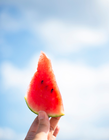 Share of watermelon in hand on blue sky with clouds. Red watermelon juicy flesh with black grains. Picnic. Summer.
