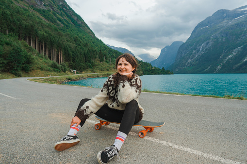 Young Caucasian woman in knitted sweater sitting on longboard near the lake in Norway