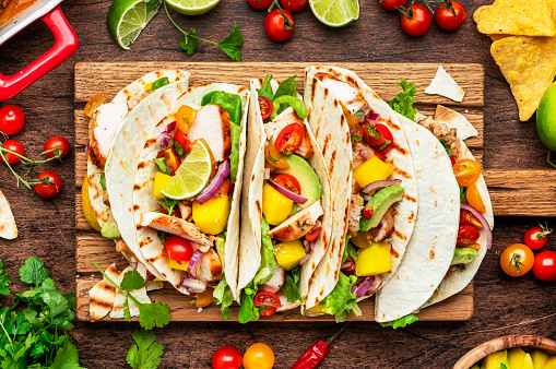 Taco party. Corn tortillas with grilled chicken fillet, salsa sauce, mango, cilantro and red onion on rustic wooden cutting board. Wood table background, top view