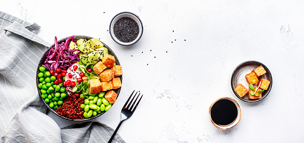 Healthy vegan food. Buddha bowl with quinoa, fried tofu, avocado, edamame, green peas, radish, cabbage and sesame seeds. White kitchen table background, top view banner