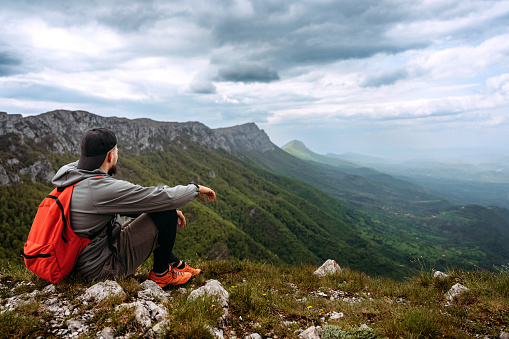 Male hiker with backpack looking at beautiful landscape scenery while sitting on stone at mountain top