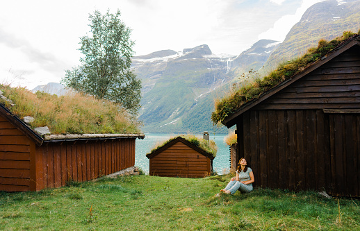 Smiling female explorer with long hair, wearing stripe shirt and orange shorts walking between old viking houses with grass on roof on the fresh green meadow in Loen, Western Norway, Scandinavia