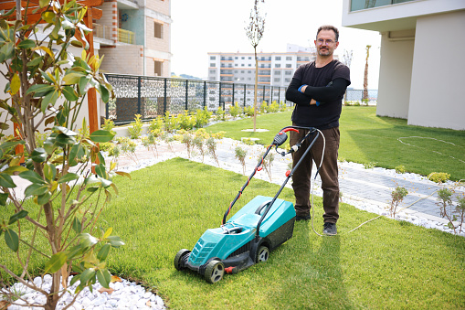 Man using a lawn mower in his back yard. Caucasian Garden and Landscaping Worker Mowing Backyard Lawn Using Electric Cordless Grass Mower. Garden Maintenance Theme.