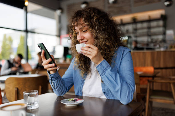 szczęśliwa młoda kobieta korzystająca z telefonu komórkowego i pijąca kawę w kawiarni - young adult lifestyles city life drinking zdjęcia i obrazy z banku zdjęć