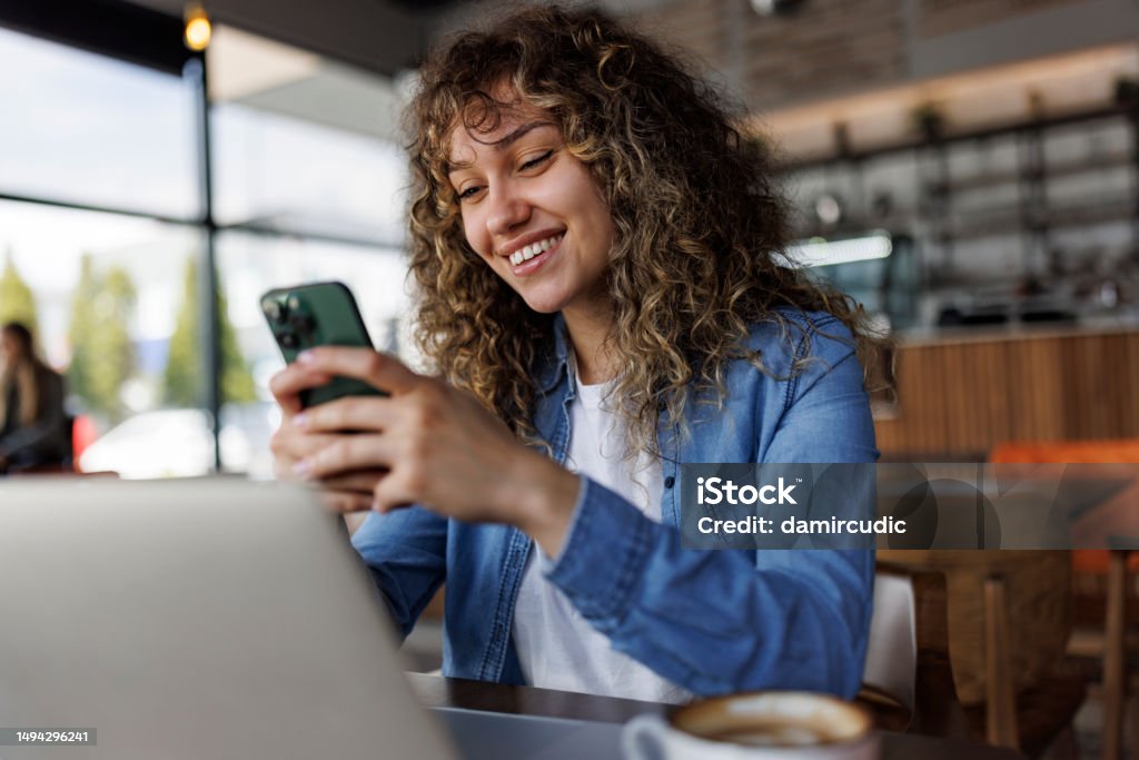 Young smiling woman using mobile phone while working on a laptop at a cafe Mobile Phone Stock Photo