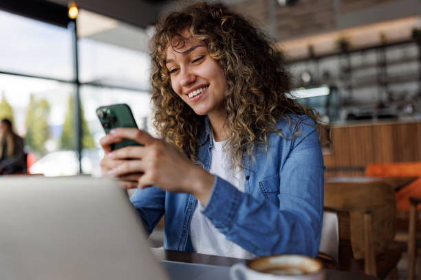 Young smiling woman using mobile phone while working on a laptop at a cafe stock photo