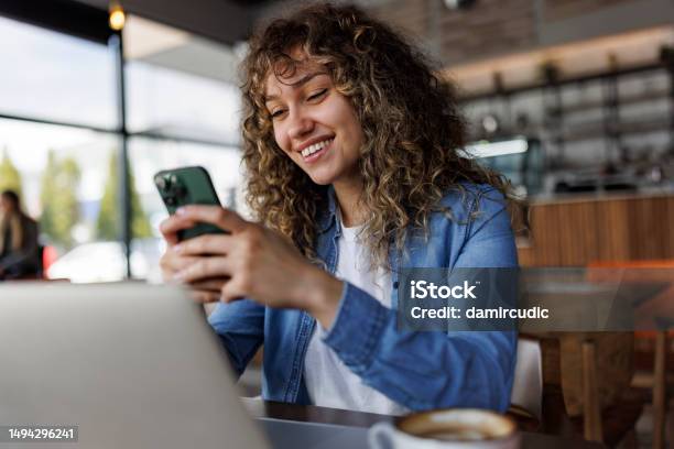 Young Smiling Woman Using Mobile Phone While Working On A Laptop At A Cafe Stock Photo - Download Image Now
