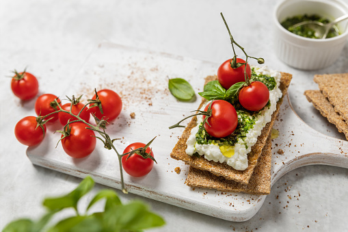 Whole grain crispbread with cottage cheese spread, cherry tomatoes and basil pesto, homemade snack or appetizer on wooden cutting board
