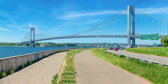 High resolution stitched panorama of the Verrazano-Narrows Bridge on a spring morning with Cars driving on the Belt Parkway. The bridge connects boroughs of Brooklyn and Staten Island in New York City. The bridge was built in 1964 and is the largest suspension bridge in the USA. Historic Fort Wadsworth is under the bridge. The photo was taken from the Shore promenade in Bay Ridge Brooklyn. Canon EOS 6D full frame censor camera. Canon EF 85mm f/1.8 USM Prime Lens. 2:1 Image Aspect Ratio. This image was downsized to 50MP. Original image resolution is 68.8MP or 11728 x 5864 px.