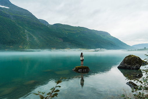 Mysterious view of woman in knitted sweater standing on the rock in the lake covered with fog