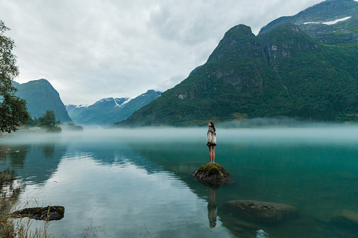 Mysterious view of woman in knitted sweater standing on the rock in the lake covered with fog