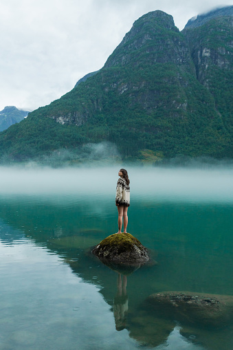 Mysterious view of woman in knitted sweater standing on the rock in the lake covered with fog