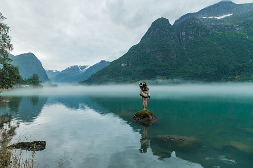 Mysterious view of woman in knitted sweater standing on the rock in the lake covered with fog