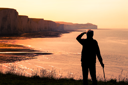 Rear view color image depicting a senior man in his 70s looking at the view of the iconic Seven Sisters cliffs on the coastline of East Sussex, UK. The man, thrown into silhouette by the setting sun, is shielding the sun from his eyes using his hand.