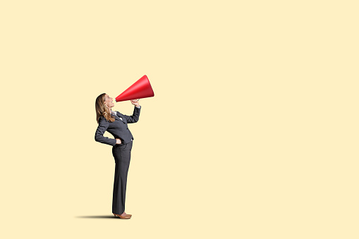 A woman standing with a hand on her hip shouts through a large red megaphone.. Isolated on a yellow background.