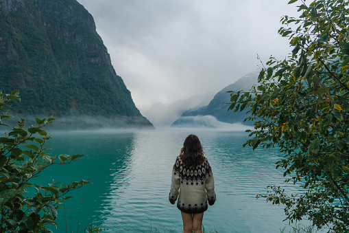 Mysterious view of woman in knitted sweater standing on the rock in the lake covered with fog