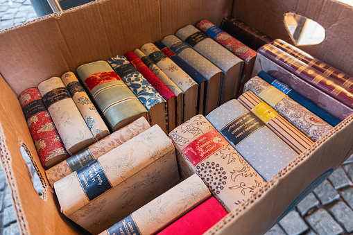 Old used italian books with cotton fabric decorated covers crammed into a cardboard box at flea market outdoors. Box full of vintage books.