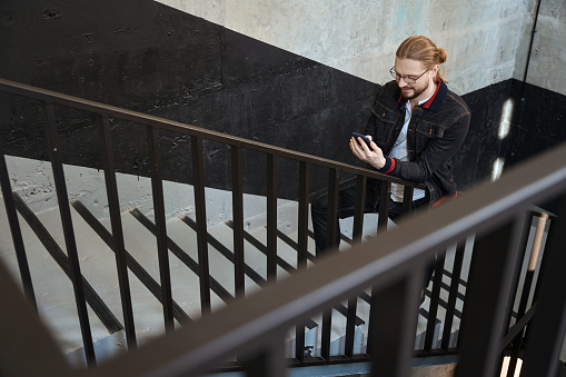 Caucasian man in fancy clothes leaning on railing and browsing mobile while walking up stair