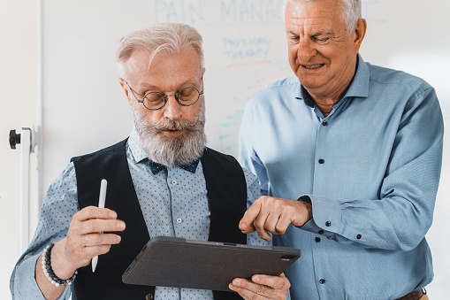 A stylish senior man with a neatly trimmed beard and bowtie holds a tablet, showing something to his colleague who's pointing at the screen. They're in an office environment, with an interactive whiteboard in the background.