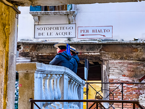 02/01/2023, Venice, Italy; Two gondoliers standing in front of From Sotoportego De Le Acque to Rialto Bridge way signs in Italian languane. Travel destination background, copy space.
