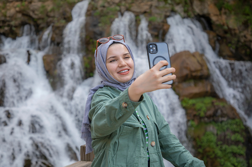 smiling young muslim woman taking selfie in front of waterfall during trip