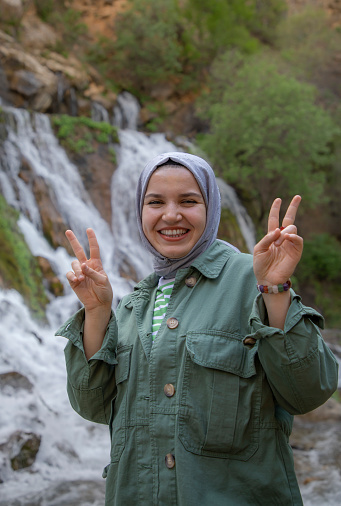 Young beautiful muslim woman smiling making peace sign in front of waterfall during trip