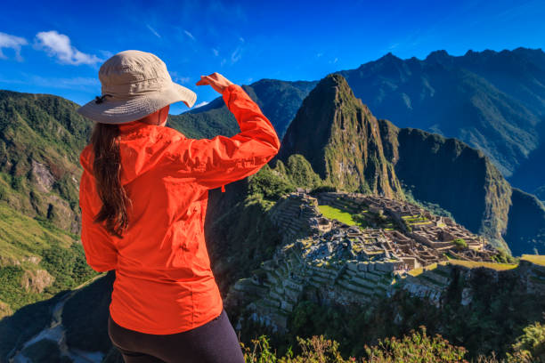 Young female tourist, wearing an orange jacket, is looking at Machu Picchu. Machu Picchu (Quechua: Machu Picchu) – \