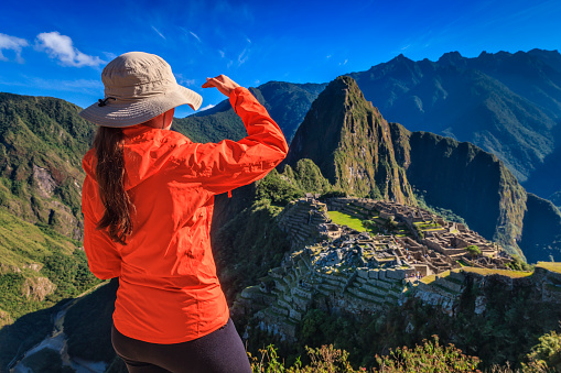 Young female tourist, wearing an orange jacket, is looking at Machu Picchu. Machu Picchu (Quechua: Machu Picchu) – 