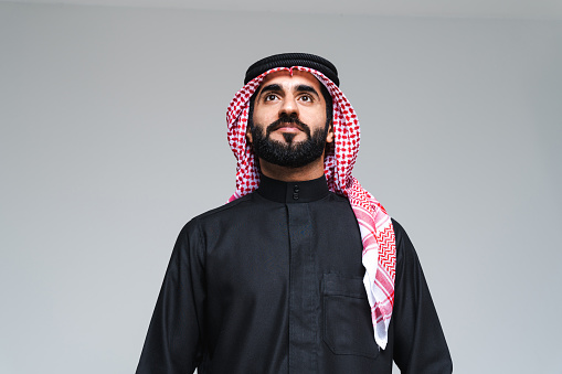 Handsome arab middle-eastern man with traditional saudi clothing in studio - Arabic muslim adult male businessman wearing thwab portrait isolated on gray background