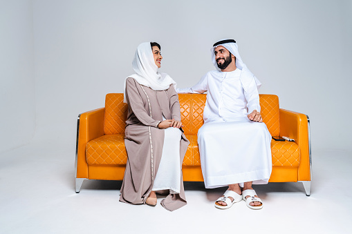 Beautiful arab middle-eastern happy couple of lovers wearing traditional abaya and kandora sitting on sofa at home- Arabic muslim adult people bonding and having fun in Dubai, United Arab Emirates