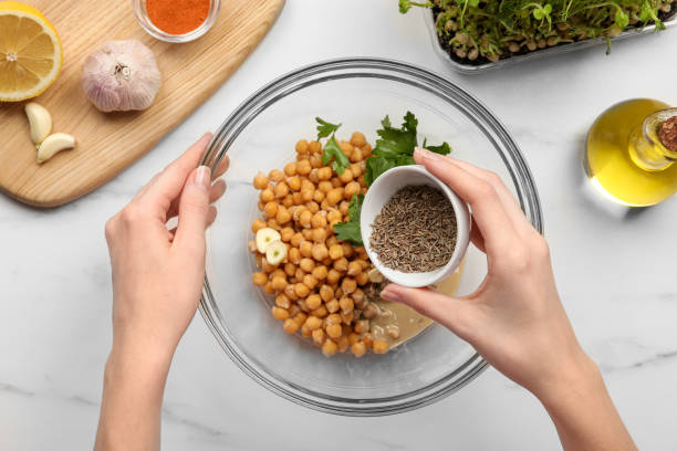 istock Woman adding cumin into bowl with ingredients for delicious hummus at white marble table, top view 1494368674