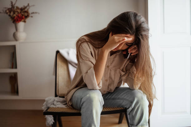 Despaired sad millennial mixed race female calls by phone, talks, sits on chair, suffering from bad news stock photo