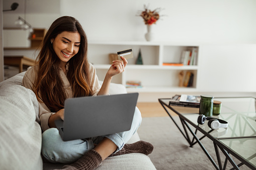 Smiling millennial mixed race woman shopaholic typing on laptop, using credit card, enjoy online shopping in living room interior. Business remotely, sale, banking and cashback at home