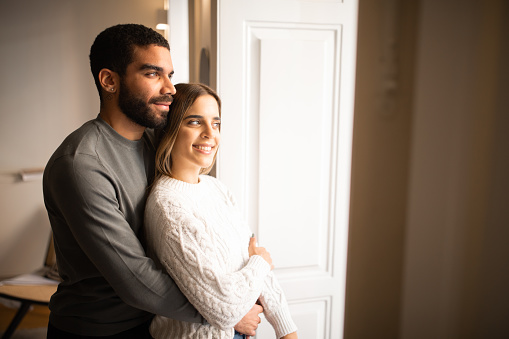 Happy multiethnic millennial couple hugging, look out the window at copy space, enjoy spare time together in minimalist living room interior. Rest at home, love and relationships, ad and offer