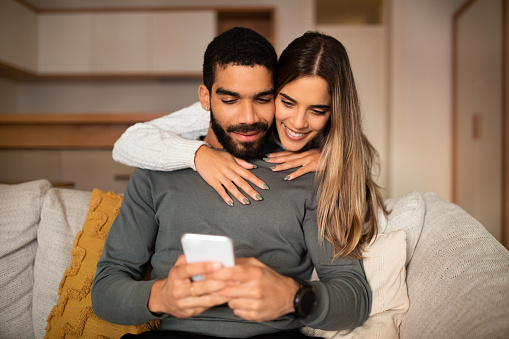 Glad young caucasian lady hugs arabic male watch video on smartphone, reads message, blog in living room interior. App and social network, communication remotely with gadget, good news