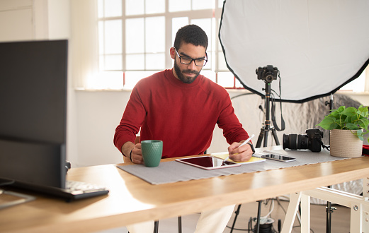 Handsome stylish young man in casual outfit sitting at desk with pc, using digital tablet, drinking coffee. Creative graphic designer professional photographer working on project at office