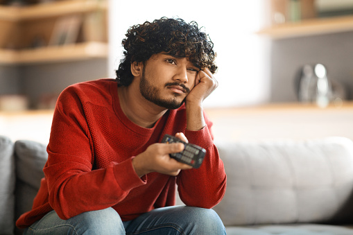 Portrait Of Bored Upset Young Indian Guy Watching TV At Home, Discontented Eastern Man Sitting On Couch At Home, Leaning Head On Hand And Switching Channels With Remote Controller, Copy Space