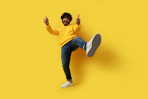 Happy handsome curly young indian man in stylish casual outfit showing thumb ups and shoe sneaker sole on yellow studio background, like something or someone, copy space. Human gestures concept