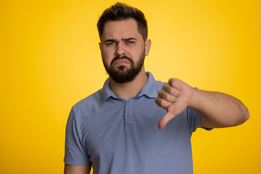 Dislike. Upset unhappy caucasian man in t-shirt showing thumbs down sign gesture, expressing discontent, disapproval, dissatisfied, dislike. Handsome young guy. Indoor studio shot on yellow background