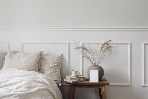 Elegant Scandinavian bedroom interior. Blank greeting card, invitation mockup on bedside table. Cup of coffee, old books. Ball vase with dry grass. Beige linen throw bedding, white wall background. elegante interior de dormitorio escandinavo. tarjeta de felicitación en blanco, maqueta de invitación en la mesita de noche. taza de café, libros viejos. jarrón de bolas con hierba seca. ropa de cama de lino beige. fondo de pared blanco.