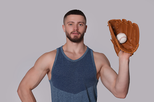 Athletic young man with glove and baseball ball on light grey background