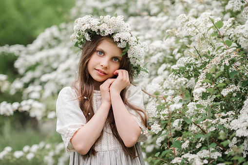 Cute stylish child girl 5-6 year old with white flowers wear floral wreath and rustic dress outdoor in garden. Springtime. Childhood.