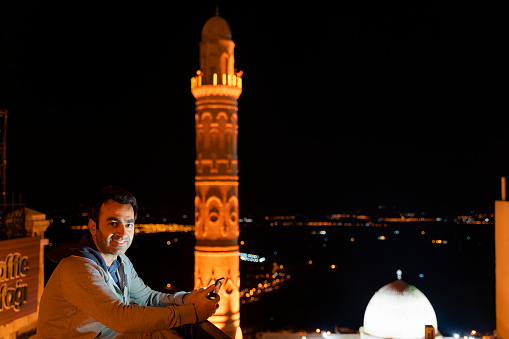 Man is using mobile phone in front of view of Mardin Grand Mosque at twilight. Ulu Mosque minaret is one of the symbolic structures of the city of Mardin, which was established on the summit of the mountain. in the back, the flat plain of Mesopotamia is seen illuminated. Taken at dusk with a full-frame camera.