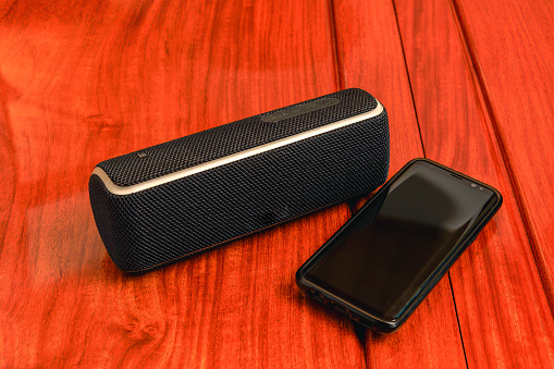 An electronic blue tooth speaker and a mobile phone are placed on a polished rosewood table. One can play music stored on the phone through the device through Blue Tooth wireless technology. Studio shot table-top photography. Copy space. Note to Inspector: Logos have been removed.