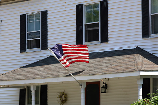 An American colonial flag with thirteeen stars representing the early colonies hanging from a wooden flagpoe in front of homes with windows and shutte