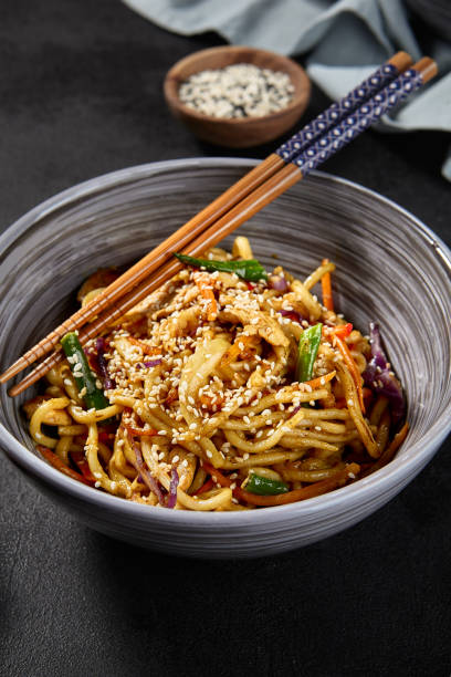 istock Wheat noodles with a variety of fresh vegetables, presented in a grey bowl, accessorized with chopsticks, on a dark backdrop. Sauce and other ingredients are scattered for additional appeal 1494440451