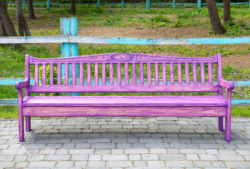 A beautiful pink wooden bench stands on a paved paving slab site in a park against the backdrop of a weathered wooden fence.