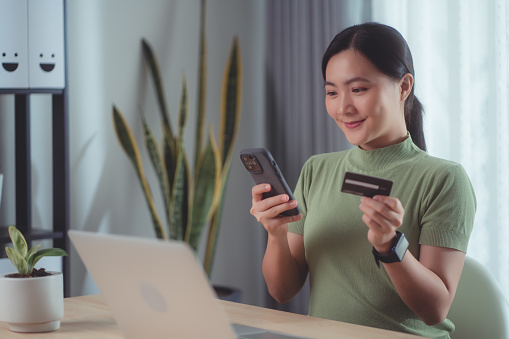 Asian woman happy with online shopping, easy online payment, holding credit card and using smartphone for shopping online at home office.